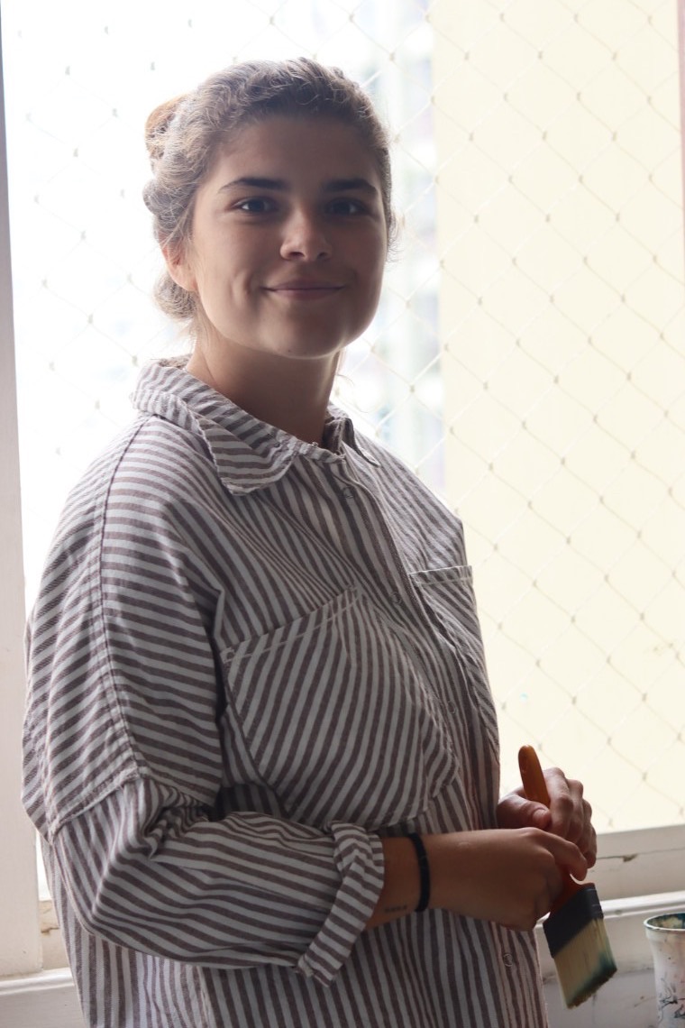 Renata Ribeiro, artista plástica, no ateliê no Rio de Janeiro, de pé na janela com o pincel na mão